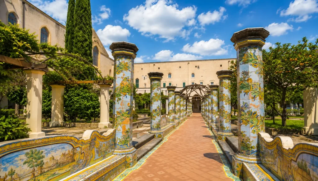 Majolica-tiled columns and peaceful garden pathways inside the Cloister of Santa Chiara in Naples under a sunny sky with soft clouds.