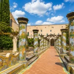 Majolica-tiled columns and peaceful garden pathways inside the Cloister of Santa Chiara in Naples under a sunny sky with soft clouds.