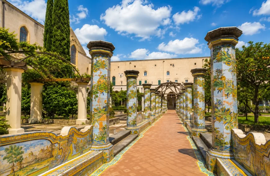 Majolica-tiled columns and peaceful garden pathways inside the Cloister of Santa Chiara in Naples under a sunny sky with soft clouds.