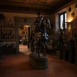 Dimly lit hall inside Museo Stibbert in Florence featuring medieval knight armor, weapons, and an armored horse display