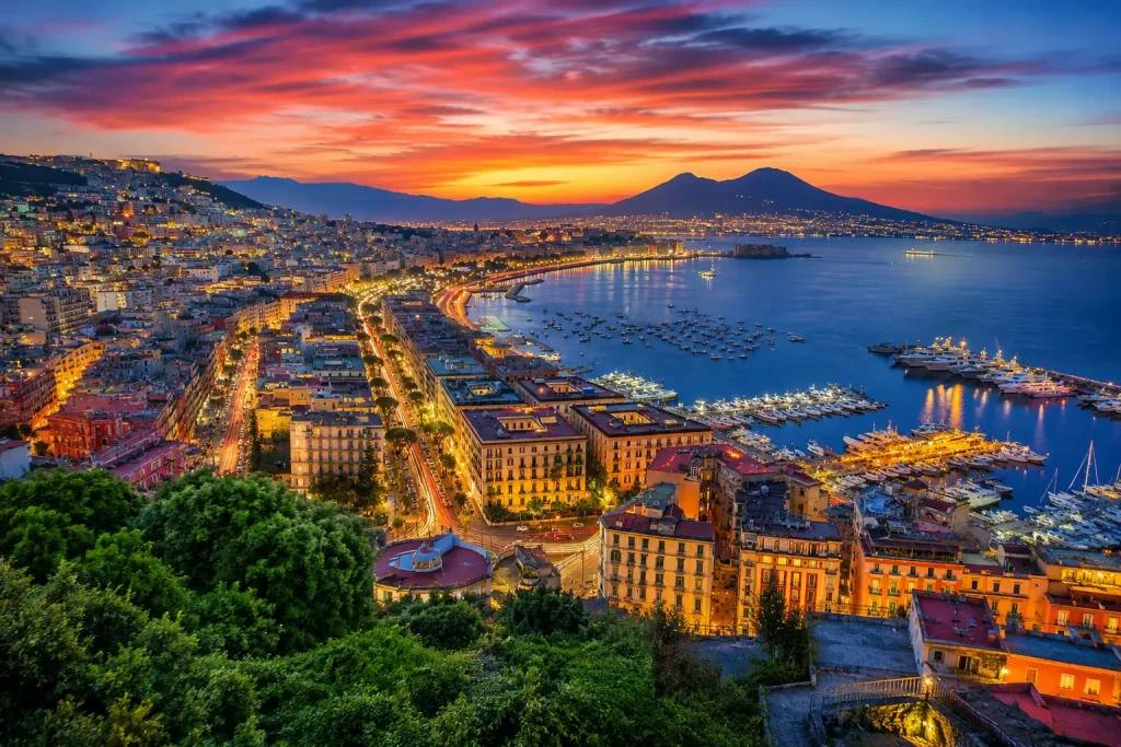 Panoramic evening view of Naples Bay with glowing city lights, marina, colorful sunset sky, and Mediterranean coastline.
