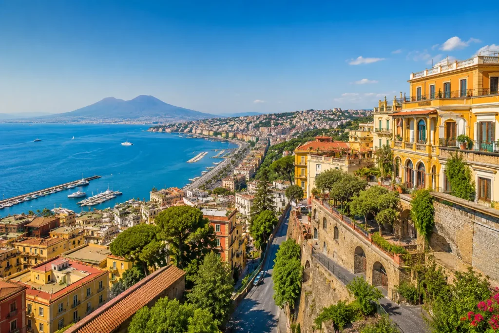 Panoramic view of Naples with Mount Vesuvius, colorful historic buildings, the Bay of Naples, and hidden hillside streets under a clear blue sky.