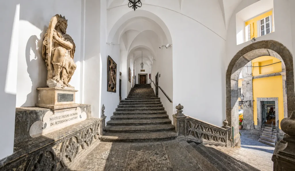 Historic staircase corridor inside Pio Monte della Misericordia in Naples with vaulted arches, stone steps, sculpture, and warm sunlight entering from the courtyard.
