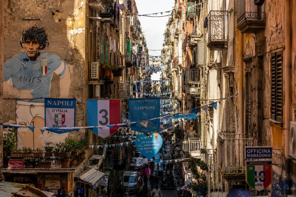 Narrow street in Quartieri Spagnoli, Naples, featuring colorful buildings, Maradona murals, hanging laundry, and authentic Neapolitan street life.