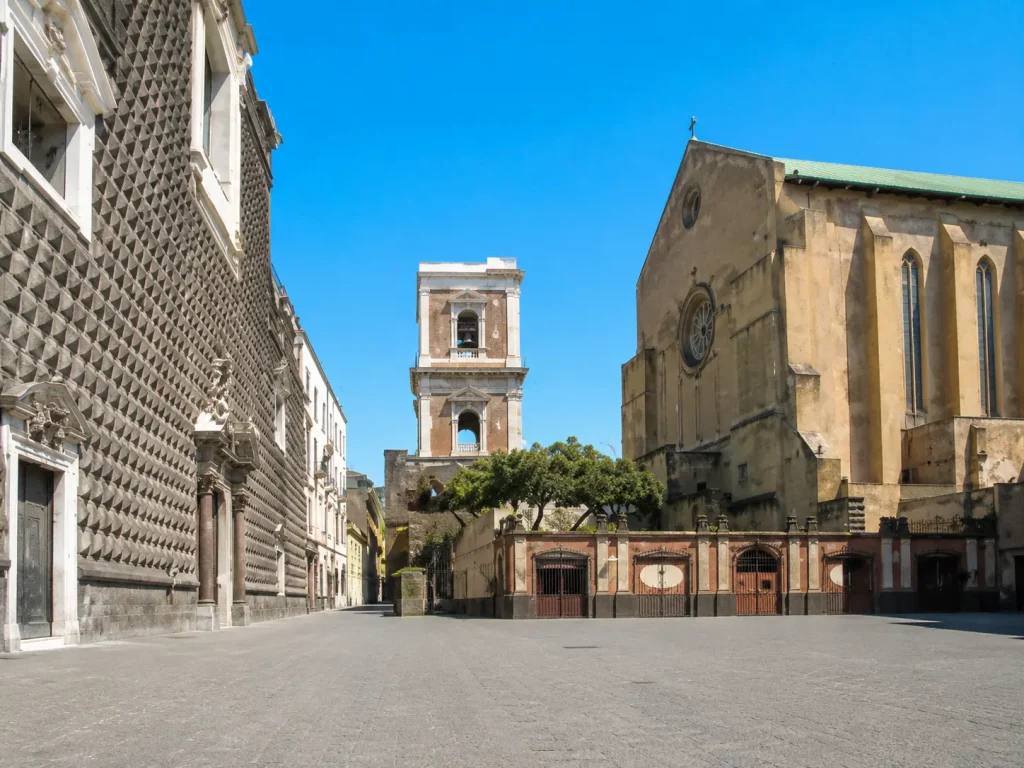 Sunny view of the Basilica and Bell Tower of Santa Chiara in Naples, seen from Piazza del Gesù Nuovo with historic architecture and an empty square.