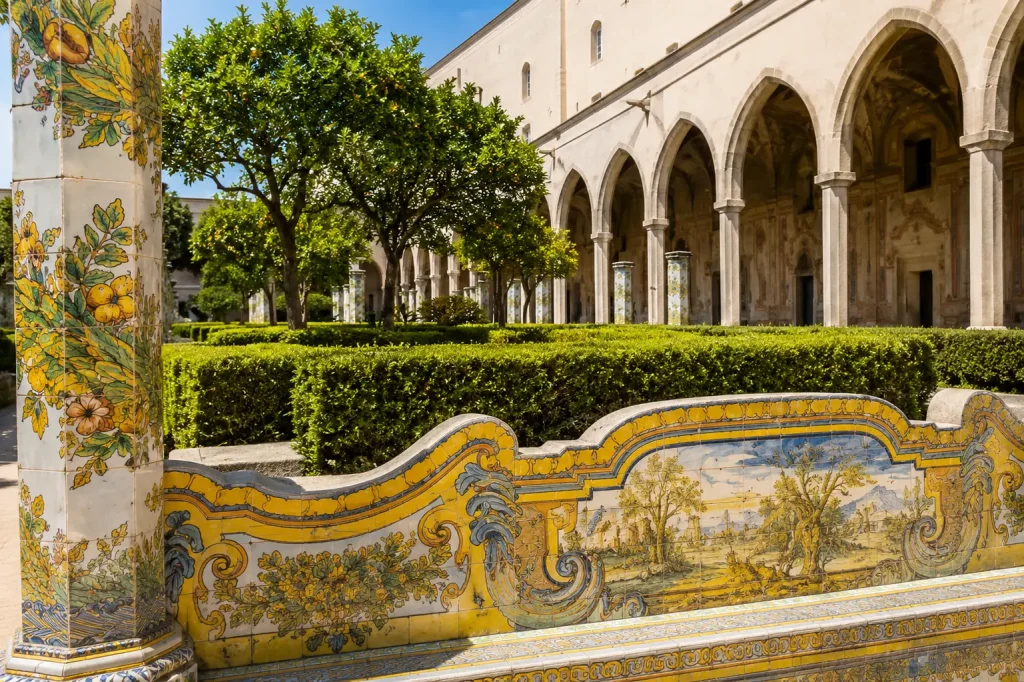 Hand-painted majolica bench and ceramic column inside the Cloister of Santa Chiara in Naples, surrounded by manicured gardens, citrus trees, and Gothic arcades on a sunny day.