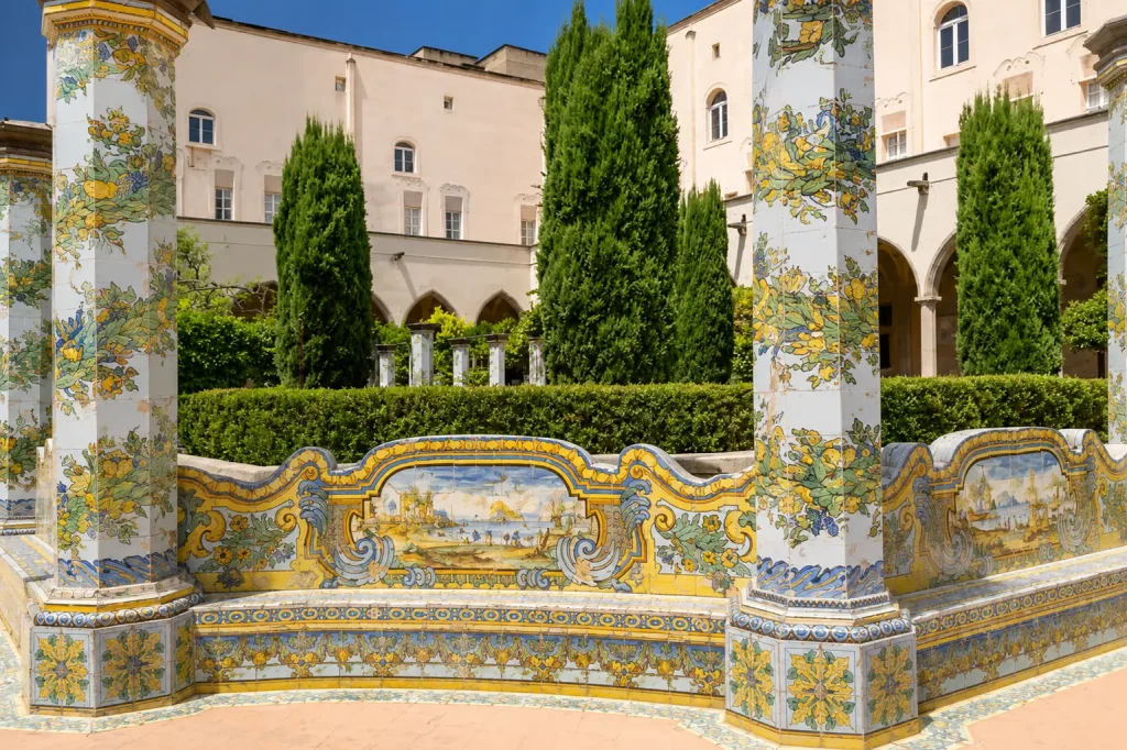 Decorative majolica-tiled bench and ceramic columns inside the Cloister of Santa Chiara in Naples, featuring colorful hand-painted scenes, floral motifs, and historic Mediterranean artwork.