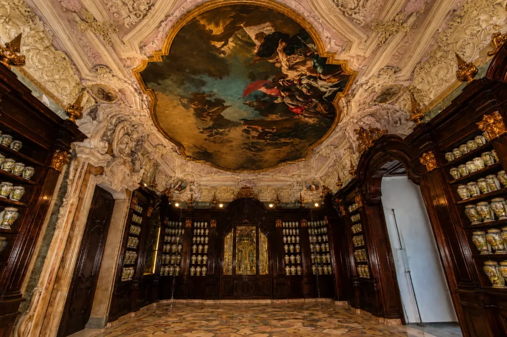 Magnificent ceiling fresco by Pietro Bardellino inside the Incurabili Pharmacy in Naples, surrounded by Baroque stucco decorations, walnut cabinets, and historic majolica apothecary jars.