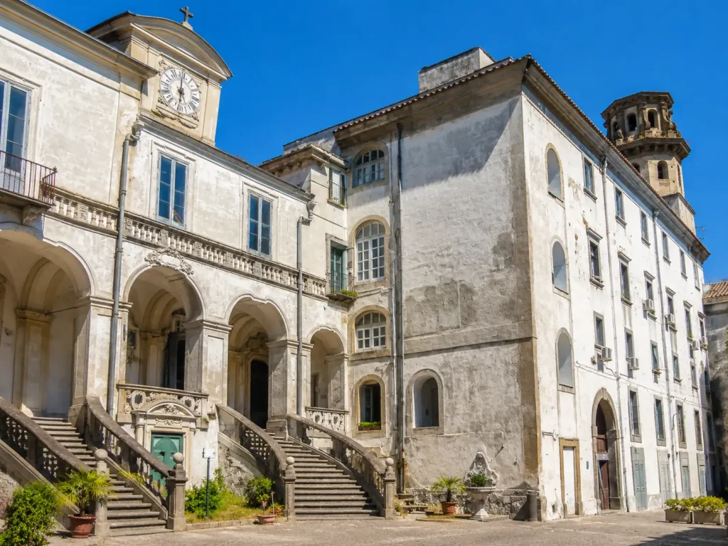 Exterior view of the historic Incurabili Pharmacy complex in Naples, featuring Baroque staircases, arcaded galleries, and the iconic clock tower under a sunny blue sky.